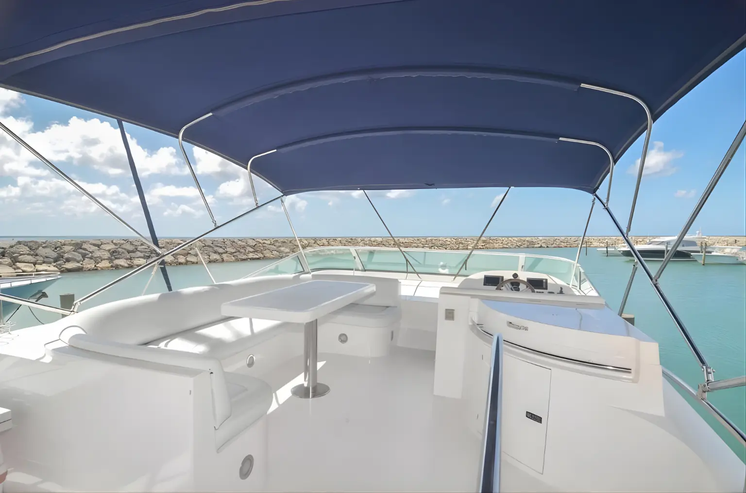Yacht flybridge with a navy bimini, white lounge seating and table, marina breakwater behind