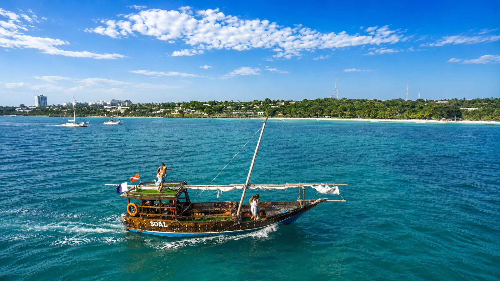 Wooden dhow cruising across turquoise water off a tree-lined Zanzibar coast with sailboats at anchor