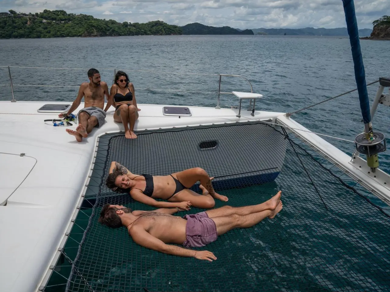 Two couples sunbathe on the bow netting and deck of the sailing catamaran in a calm green bay