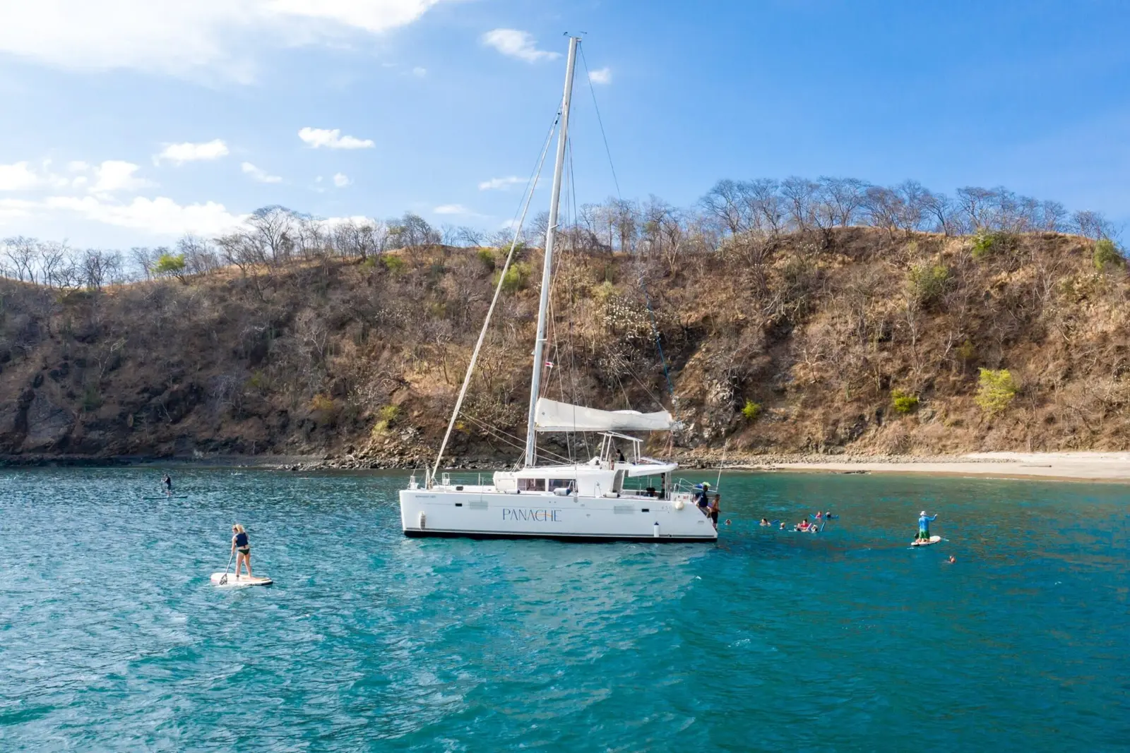 Sailing catamaran anchored in a quiet Costa Rica cove below dry forested hills as guests paddleboard nearby