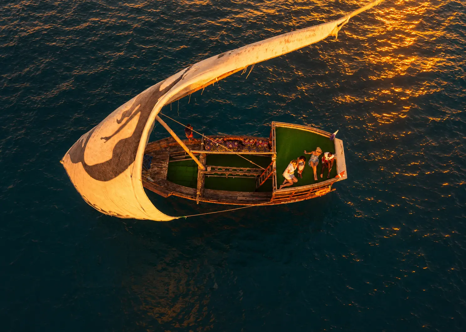 Overhead view of guests lounging on a dhow's deck under a full lateen sail on golden water off Zanzibar