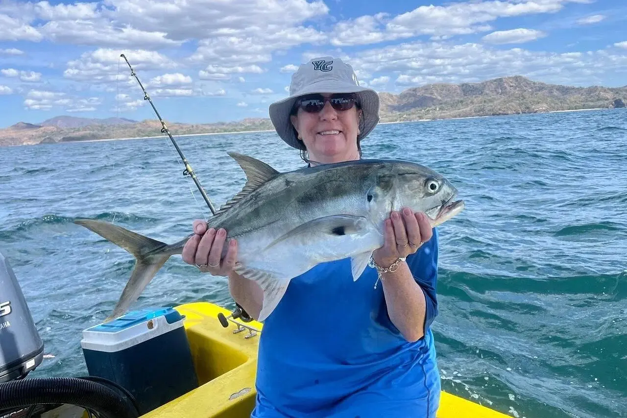 A woman in a bucket hat holds a silver jack aboard the yellow panga, dry hills behind