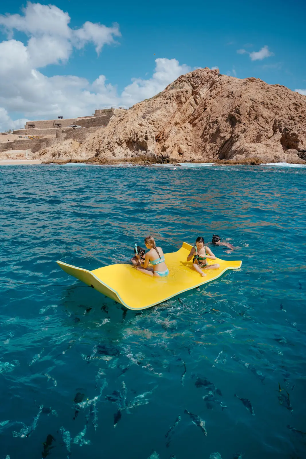 Three guests lounge on a yellow floating mat above fish near a rocky headland