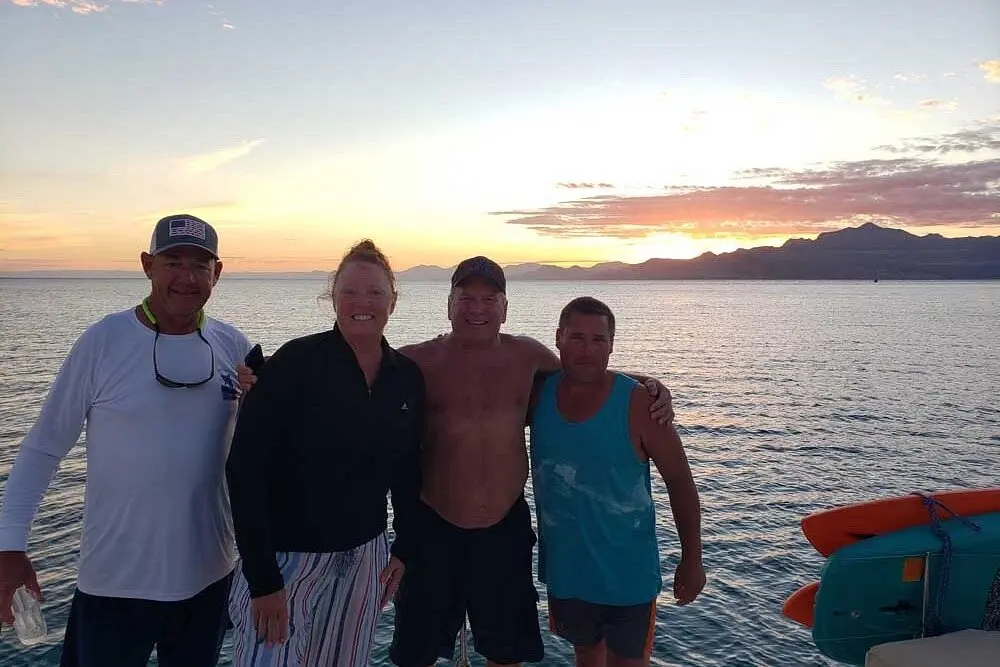 Four guests posing on a sailboat deck at sunset with calm water and Baja mountains behind