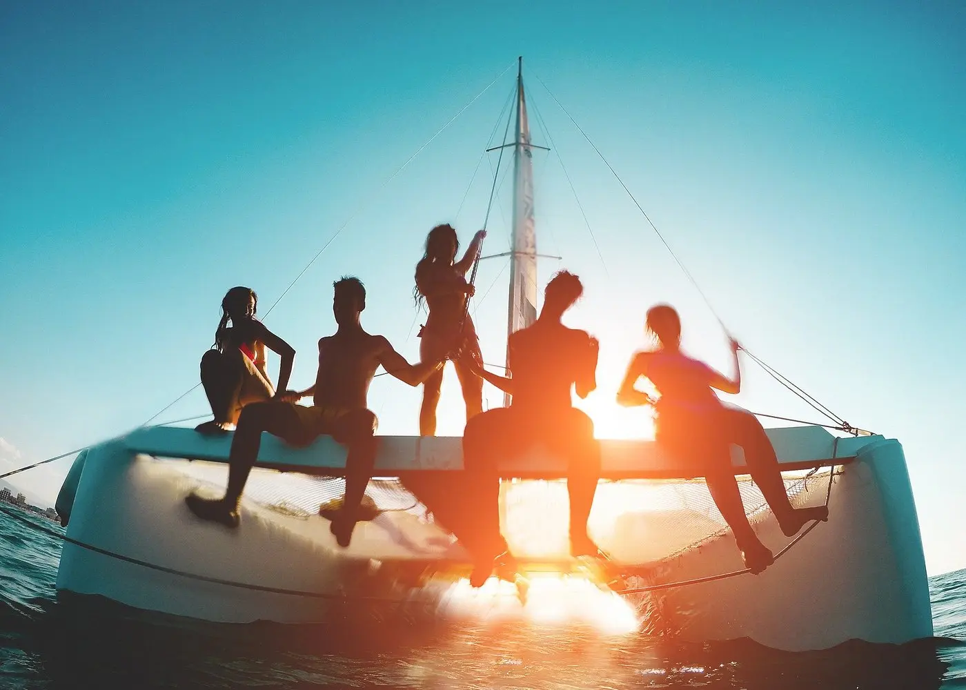 Guests silhouetted against a bright sun flare as they sit along the edge of a catamaran