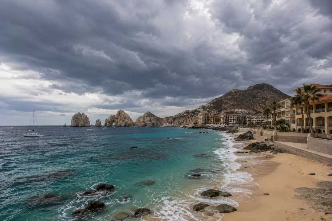 Stormy Cabo San Lucas coastline with turquoise water, sandy beach, resorts, and iconic rock formations.