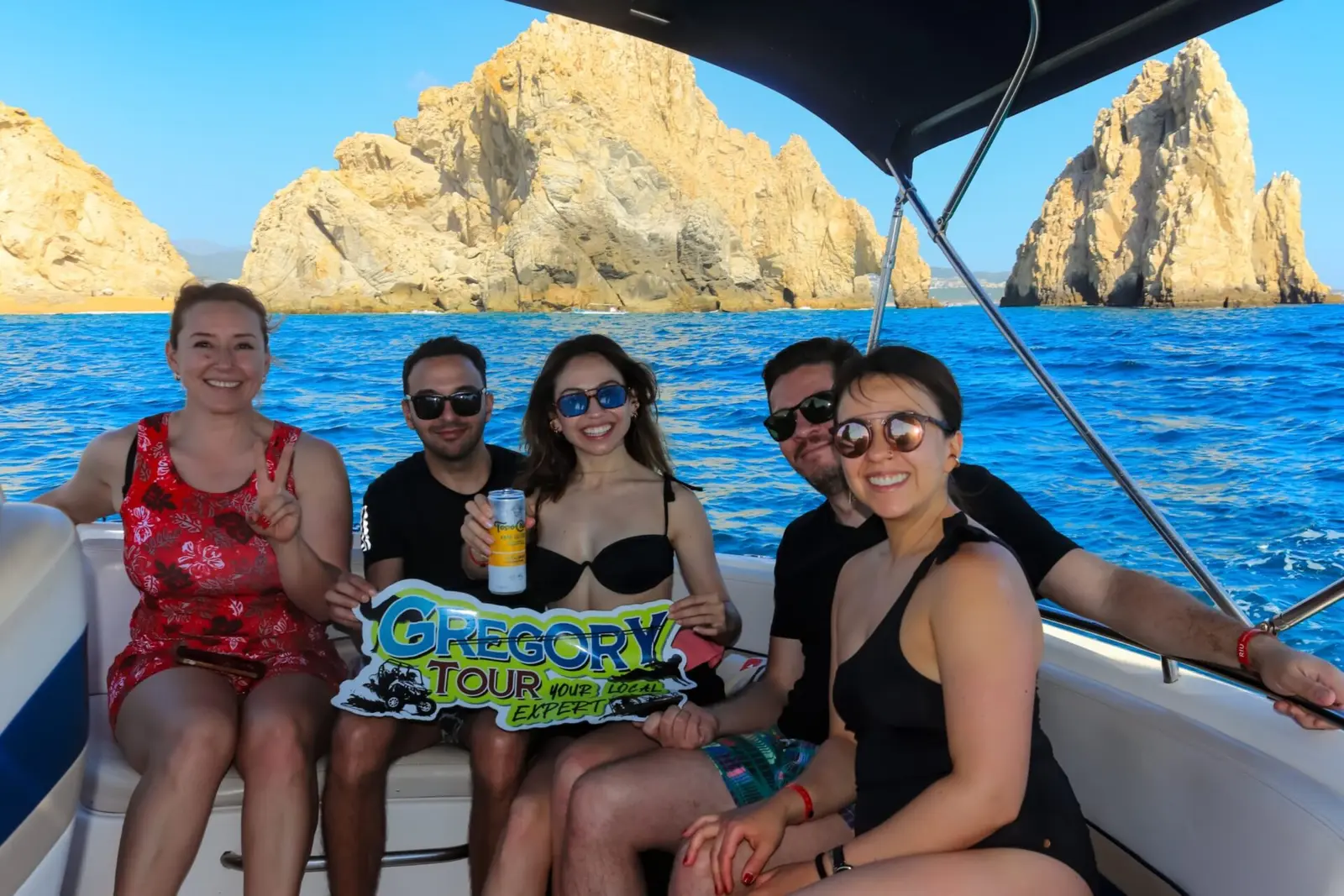 Five friends holding up a banner on a small yacht, golden Cabo San Lucas rock formations behind