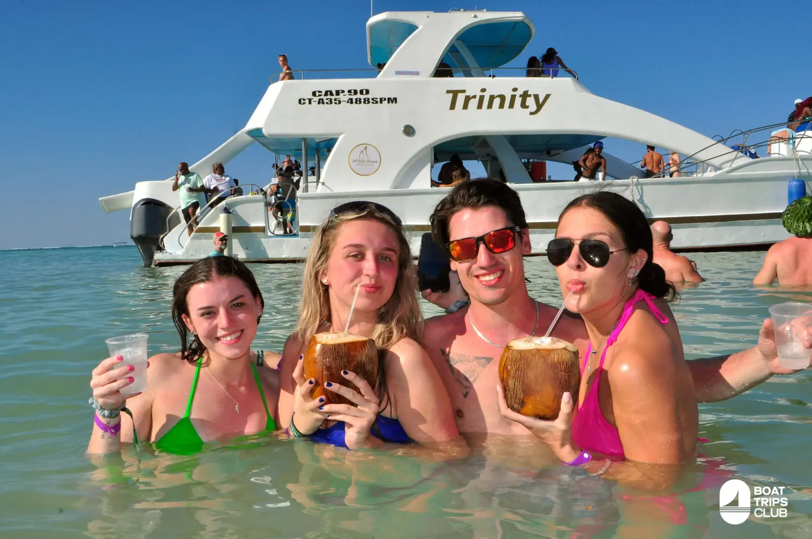 Four friends sip from coconuts in waist-deep water in front of an anchored power catamaran