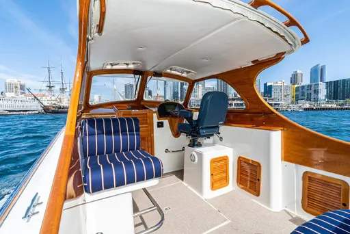 Varnished wood cockpit of a picnic boat with navy striped seating and a helm chair, a city harbor beyond
