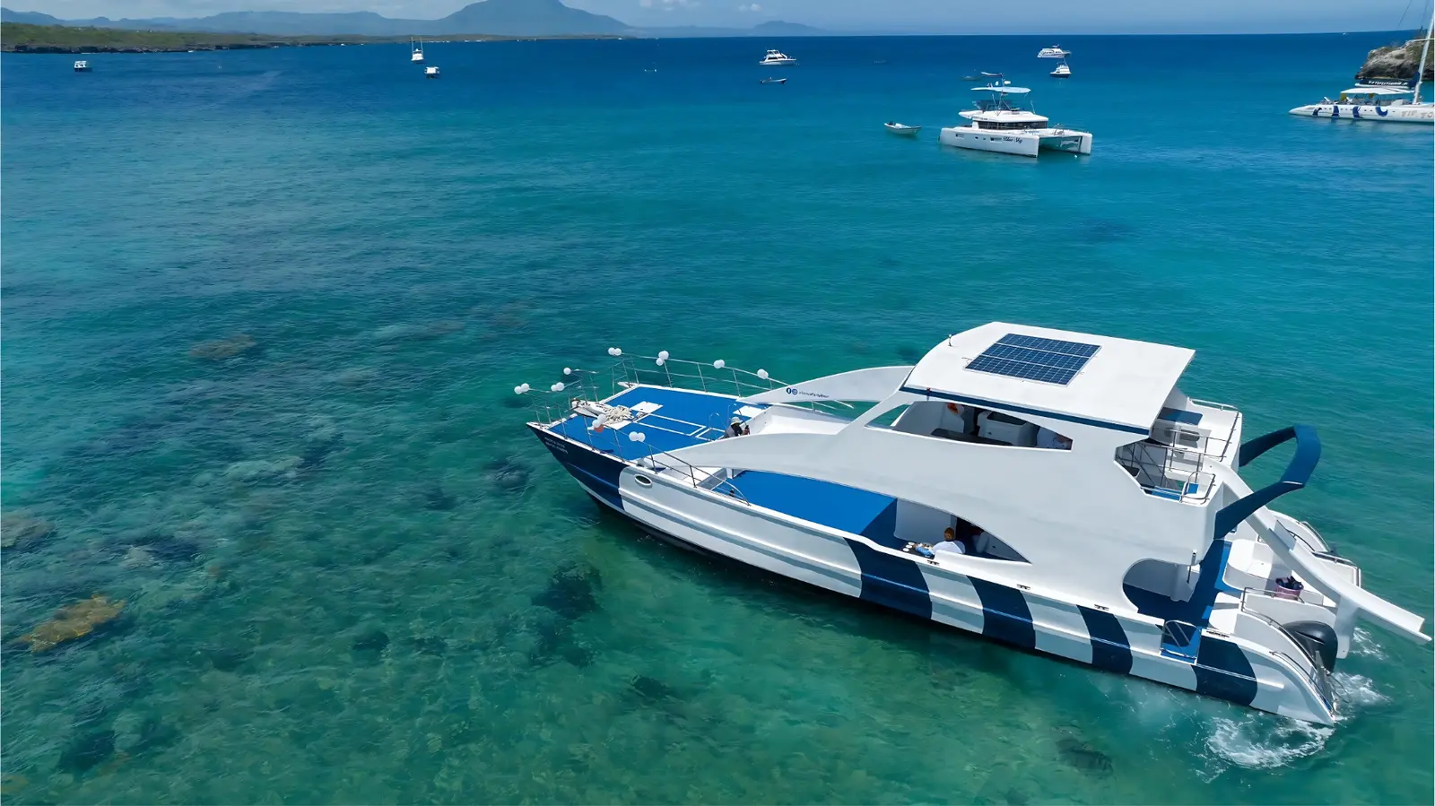 Aerial view of a catamaran with a solar-panel roof on turquoise water over a reef, boats in the distance