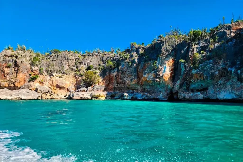 Rugged orange-streaked cliffs above turquoise water on the Jaragua coast, a boat wake in front