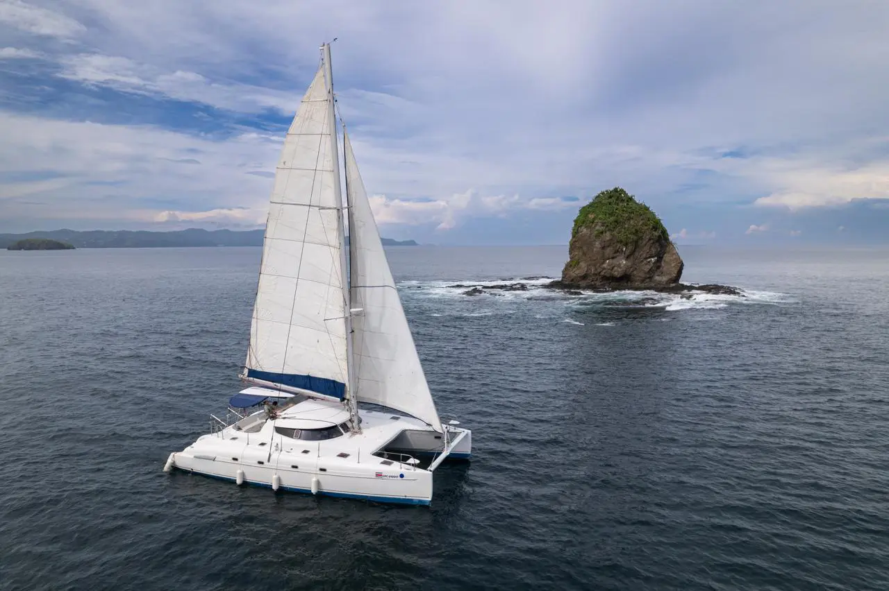 Sailing catamaran passing a tree-topped rocky islet under a cloudy sky off the Guanacaste coast