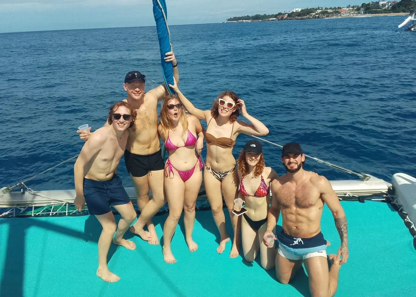 Six guests in swimwear posing on the turquoise bow net of a catamaran off Puerto Plata