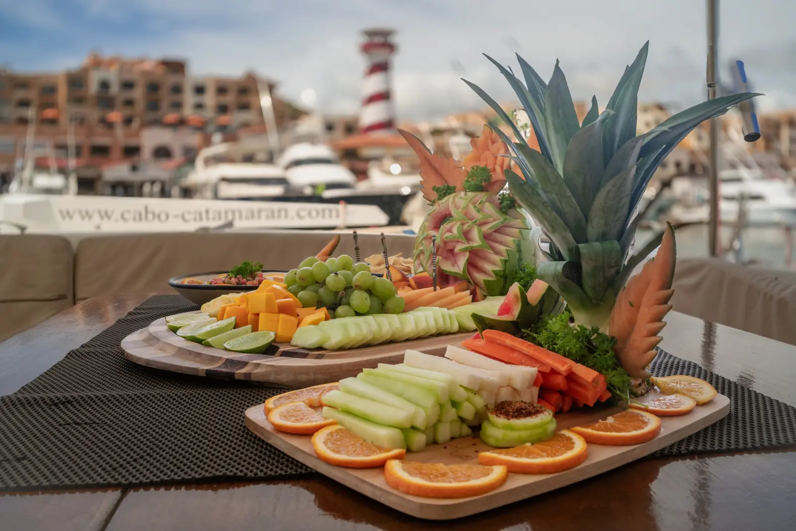 Carved fruit and vegetable platters on a catamaran table, Cabo San Lucas marina and striped lighthouse behind