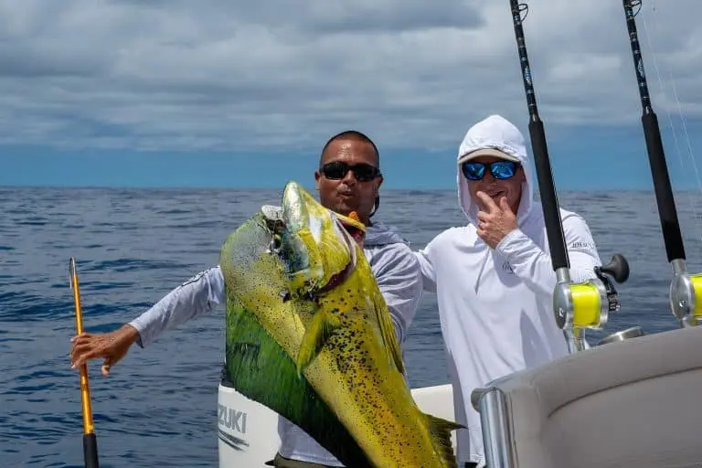 Close-up of a crew member holding up a big mahi-mahi beside a hooded angler in sunglasses