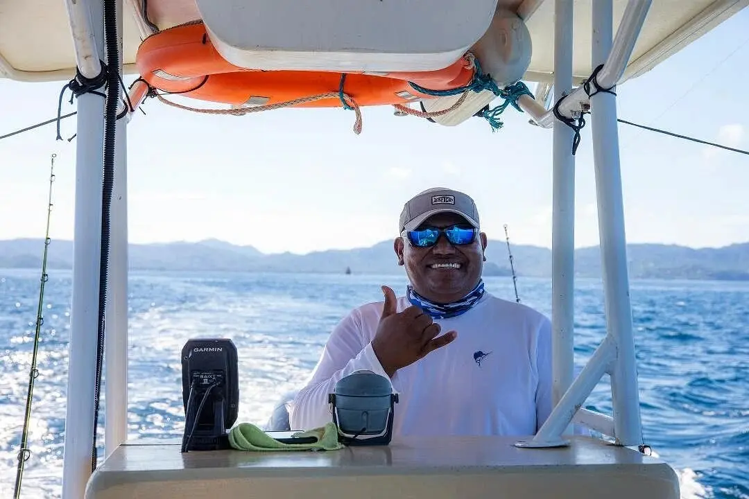 Captain at the console under the T-top flashing a shaka sign, the Playa Flamingo coast behind
