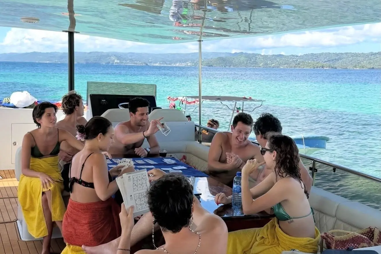 Friends playing cards around a table on the catamaran's shaded deck, green Samaná hills across the bay