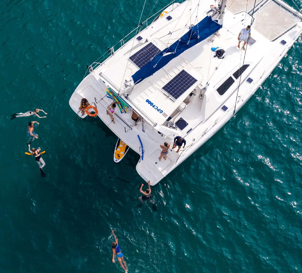 Aerial view of snorkelers swimming beside a sailing catamaran in clear teal water