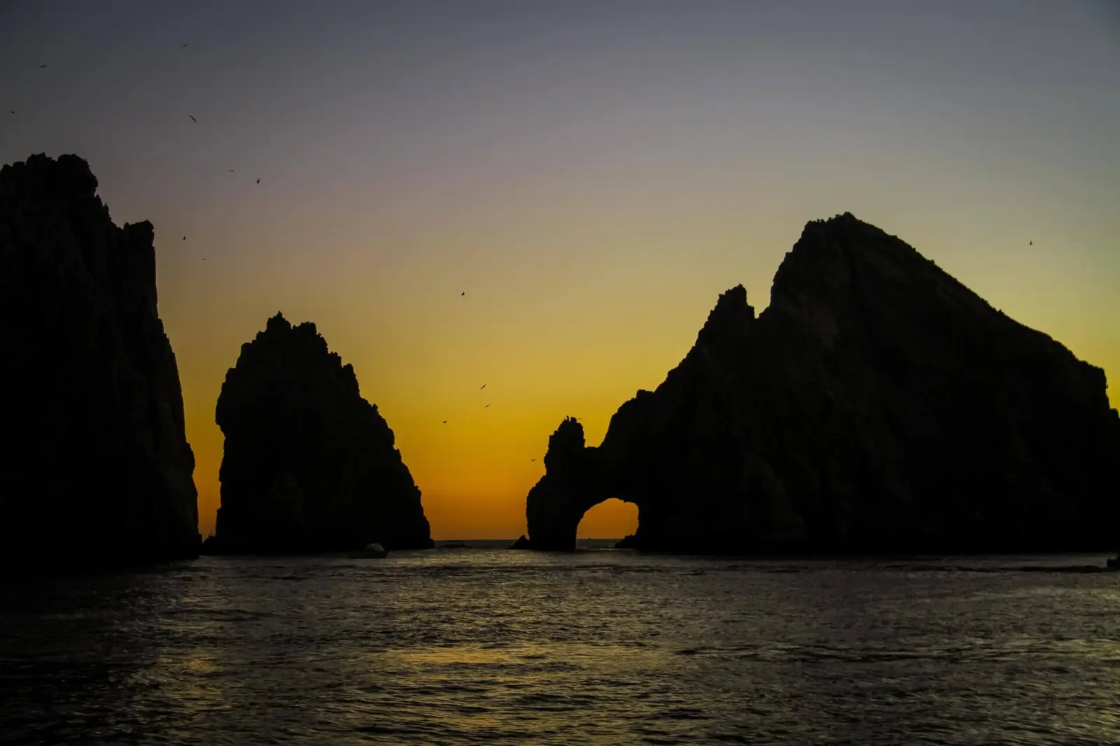 El Arco and neighboring rocks in silhouette against a golden dusk sky over calm water