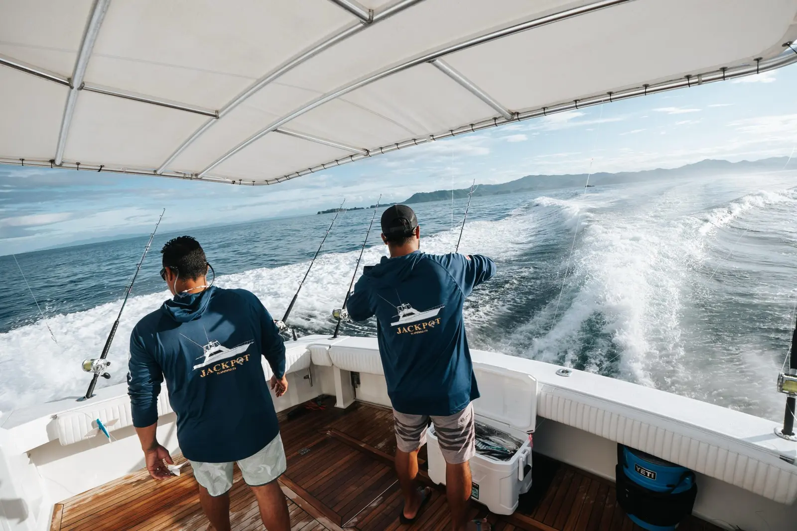 Two crew members in matching shirts watch the trolling lines from the cockpit as the yacht's wake churns