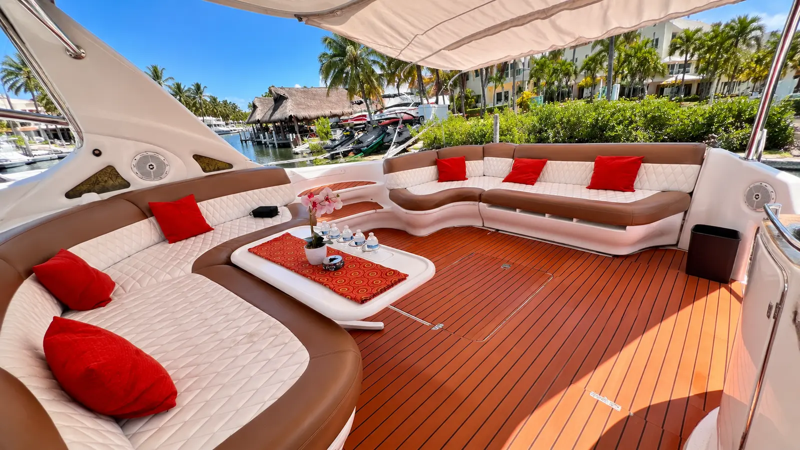 Cockpit lounge of a yacht with white sofas, red cushions and a teak floor, docked beside palm trees