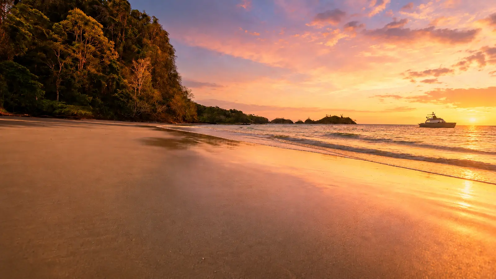 Sunset glow over an empty sandy beach and forested headland with a yacht anchored offshore in Costa Rica