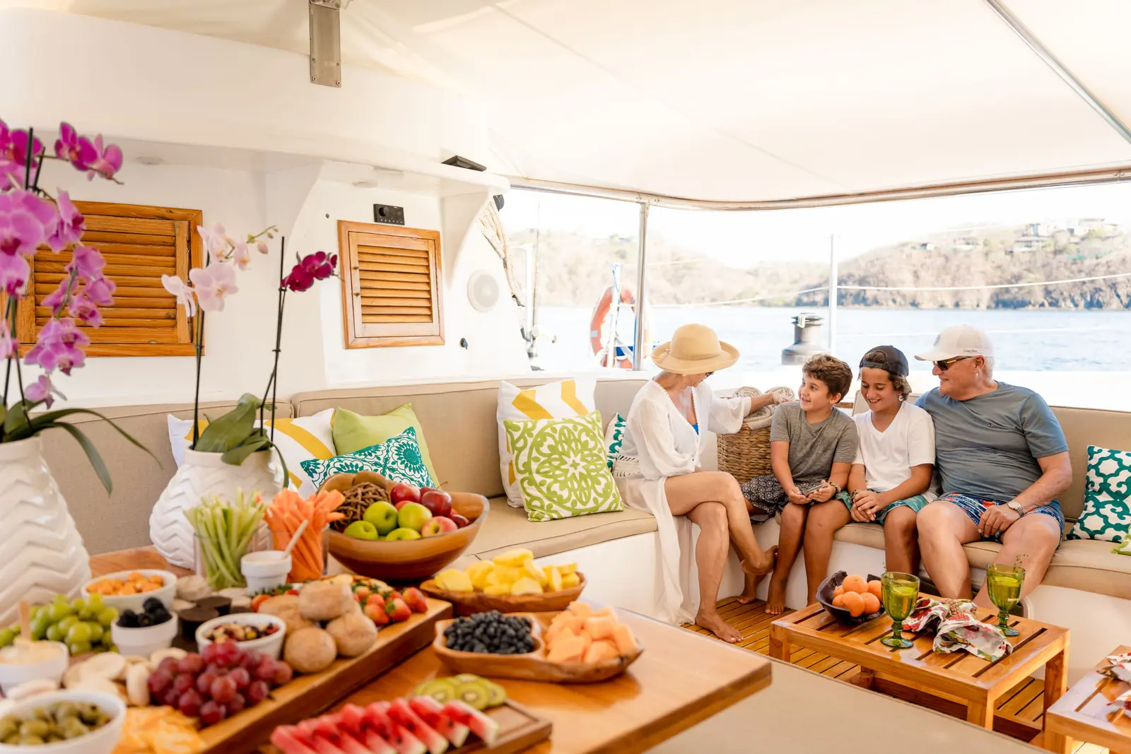 A family sits beside a spread of fruit, cheese and orchids in the catamaran lounge