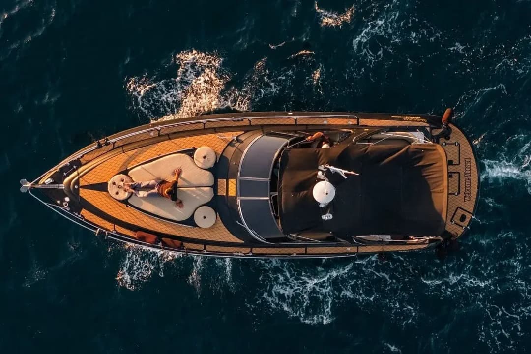 Aerial view of a motor yacht cruising dark blue water at golden hour, a guest lounging on the bow sunpad