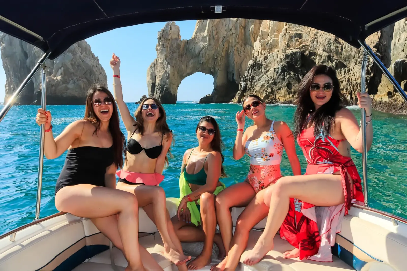 Five friends in swimsuits striking poses at the bow of a canopied yacht with El Arco behind