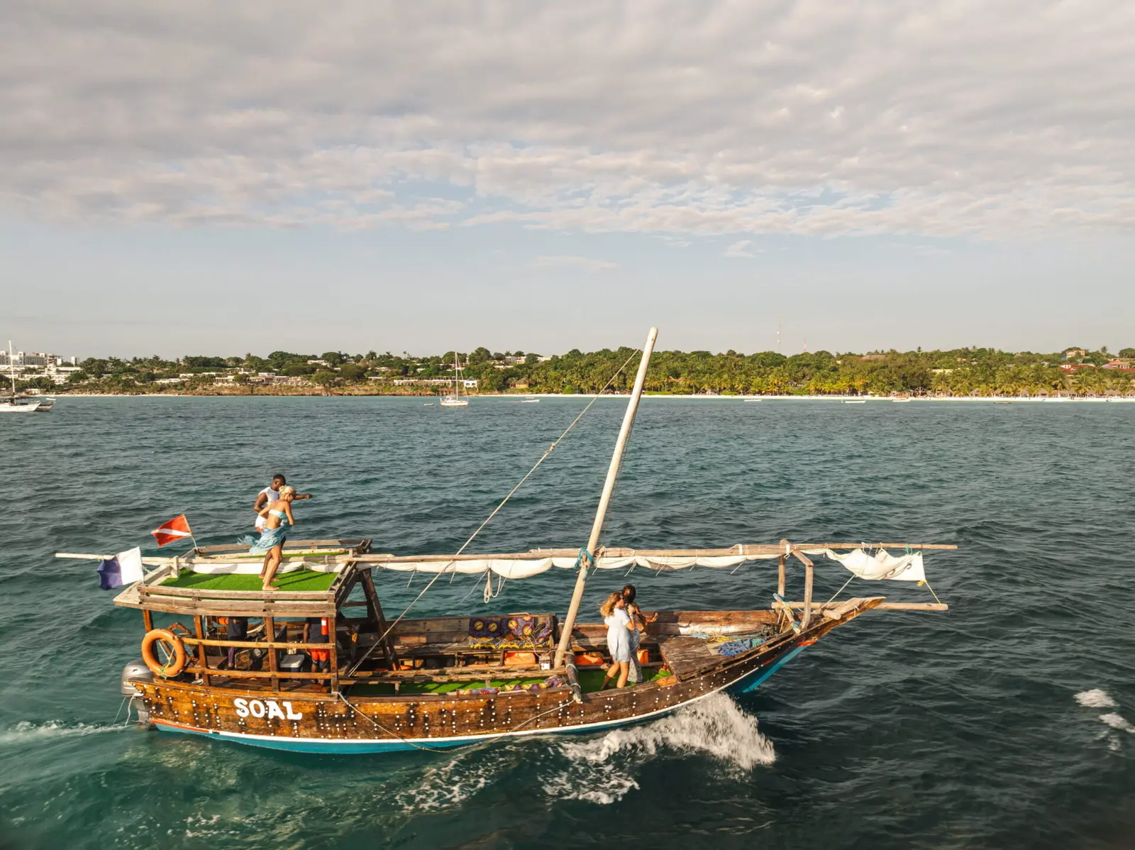 Dhow leaving a white wake as it motors past Zanzibar's green coastline, a man on the roof deck