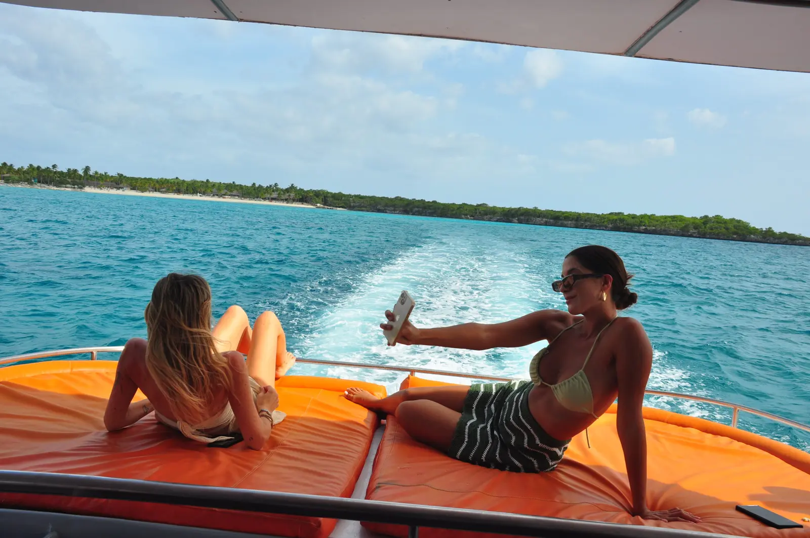 Two women taking a selfie on an orange sun pad as the power catamaran's wake trails toward the coast