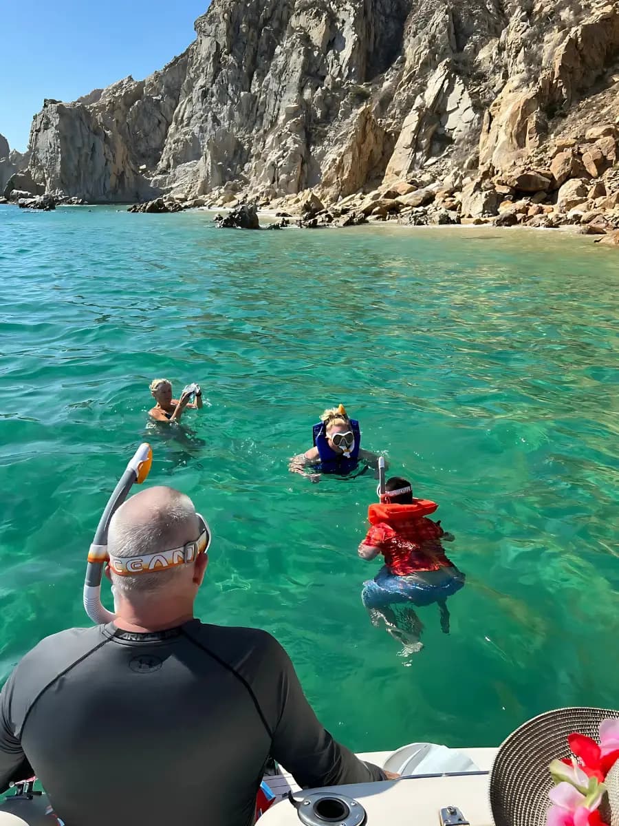 Man in a rash guard watching snorkelers swim beside the yacht below the cliffs of Cabo San Lucas