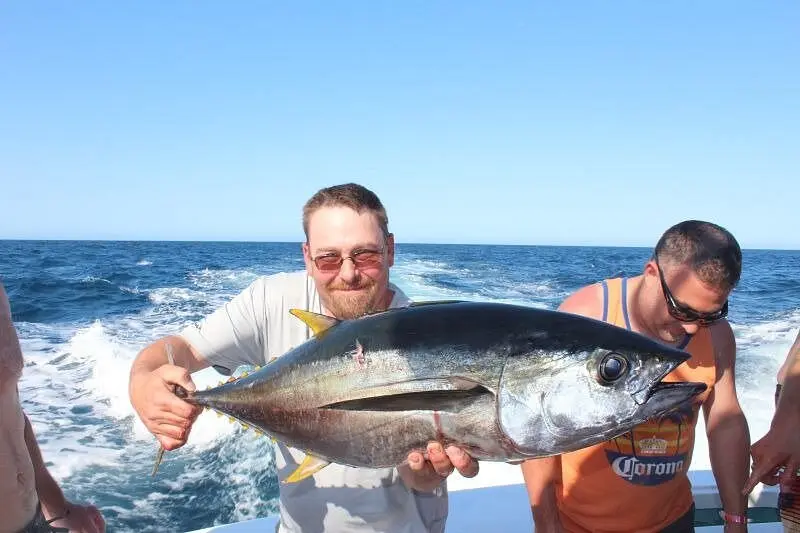 Two anglers hold up a yellowfin tuna at the boat's stern with whitewater from the wake behind