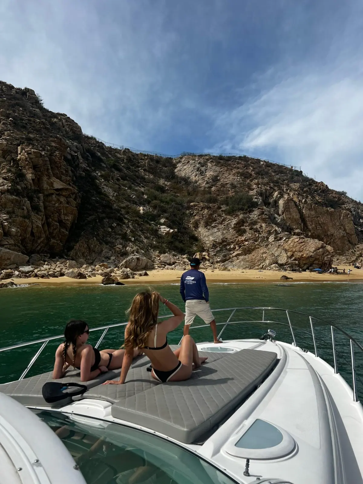Yacht anchored off a sandy Los Cabos beach below rugged brown hills, guests lounging on the bow