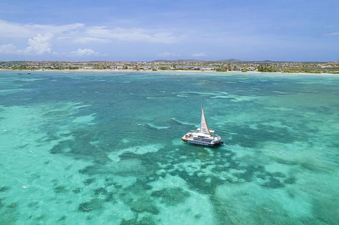 Aerial view of a sailing catamaran anchored in turquoise reef shallows off Aruba's hotel-lined coast