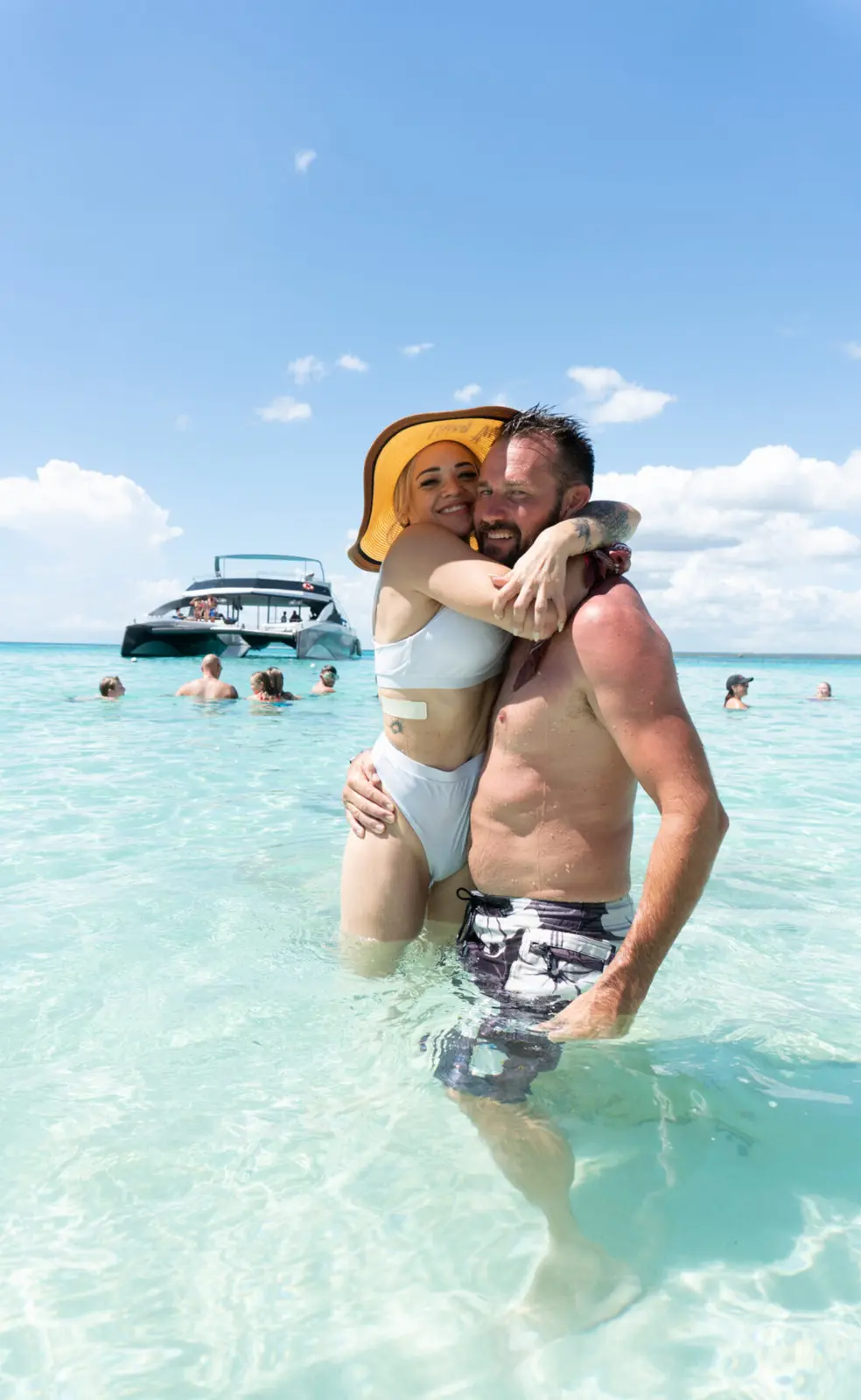Man lifting a woman in a yellow sun hat in shallow turquoise water near Punta Cana, catamaran behind