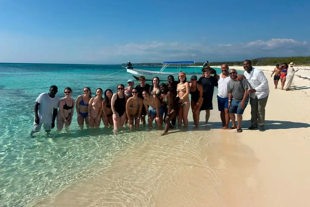 Large group of guests posing on the white sand of Bahía de las Águilas, a catamaran anchored behind