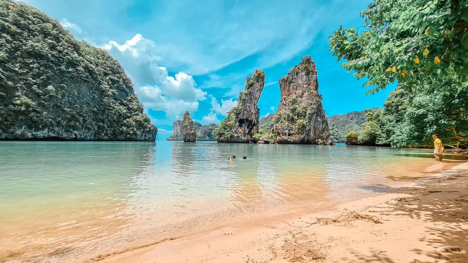 Sandy beach on calm turquoise water facing tall limestone pinnacles in Phang Nga Bay