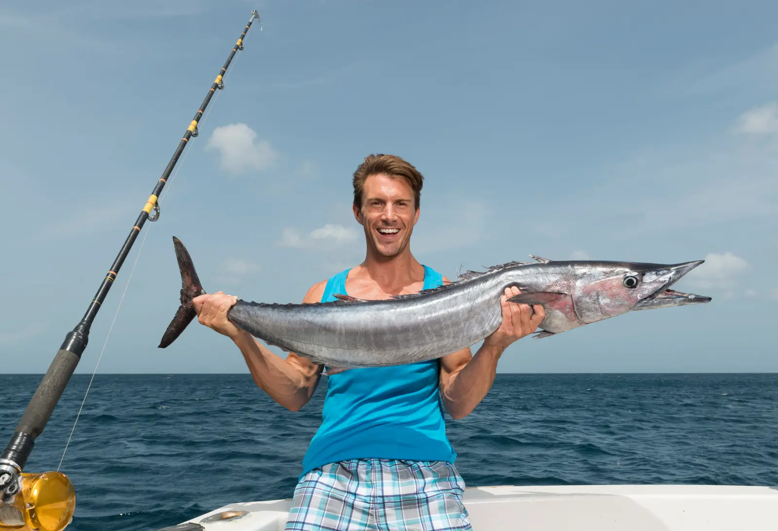 Man in a blue tank top grinning as he holds up a long silver wahoo at sea, a heavy rod beside him
