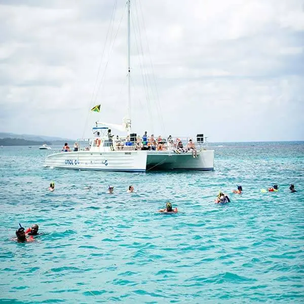 Snorkelers swimming in turquoise water in front of an anchored sailing catamaran off Ocho Rios