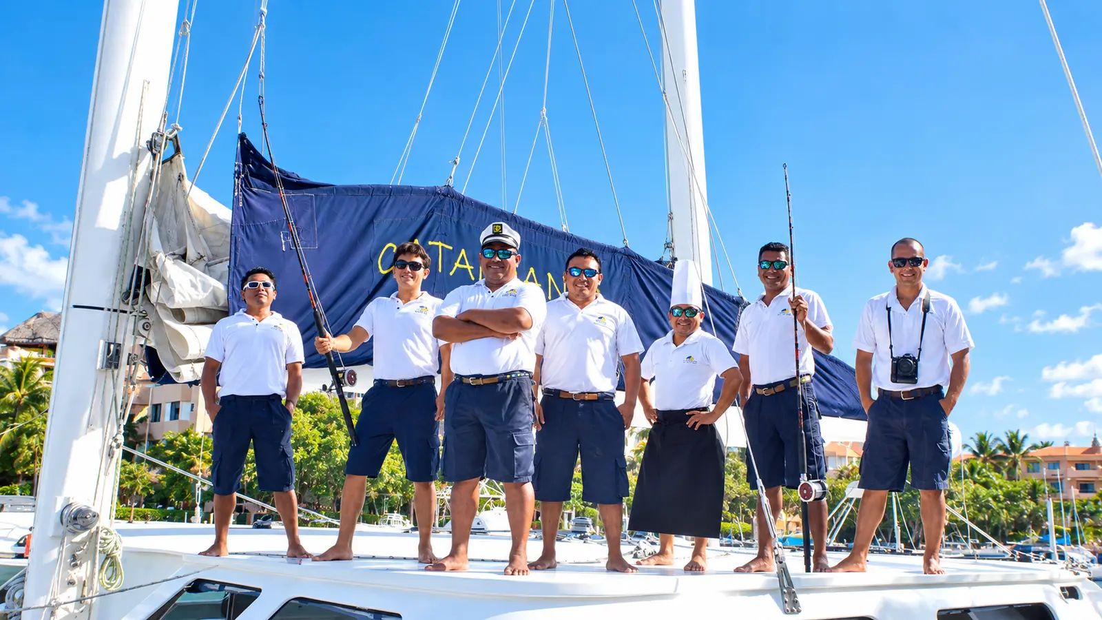 Crew in white shirts and navy shorts lined up on the sailing catamaran's deck beneath the furled sail