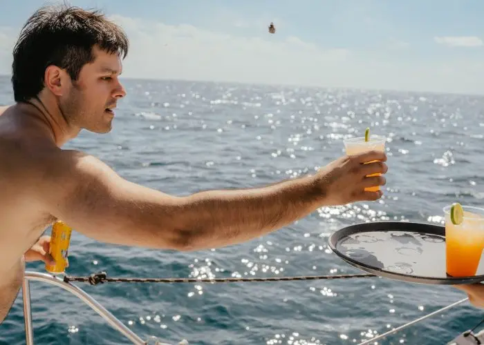 Shirtless man reaching for a cocktail from a tray held out at the rail, sun glinting on the sea