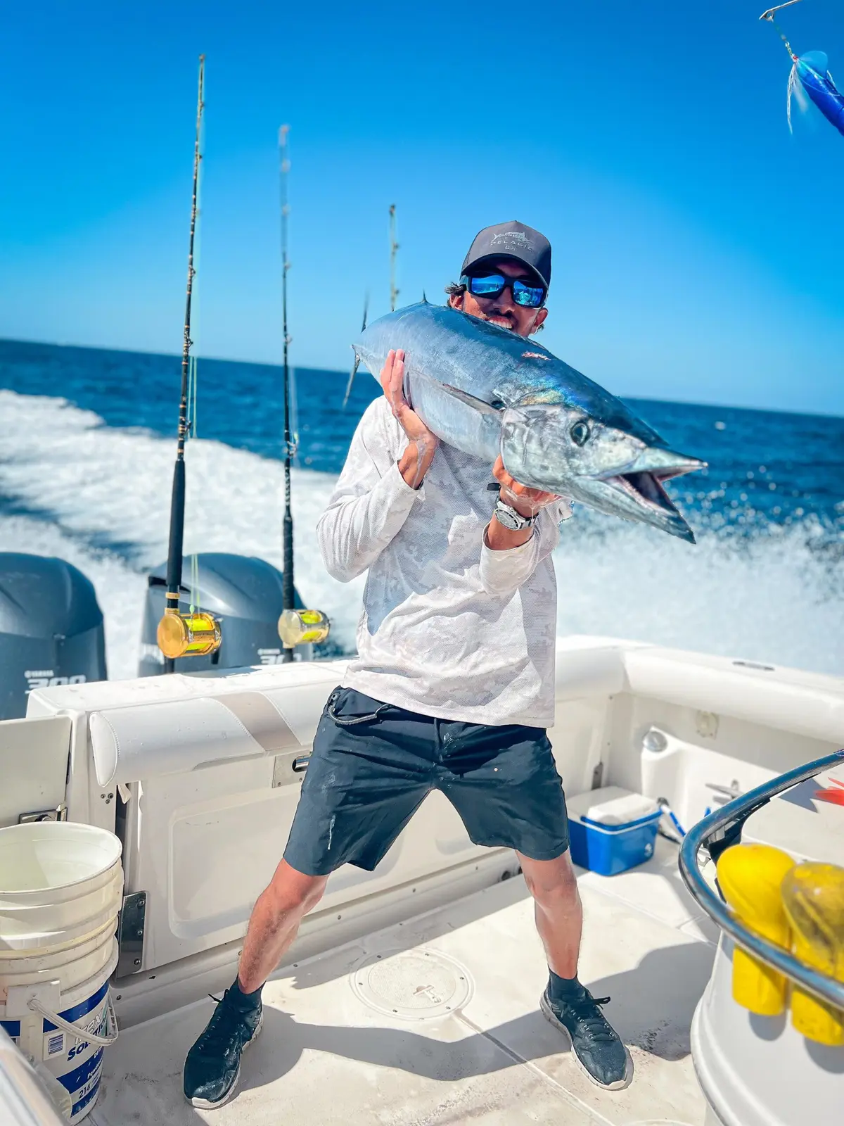 Man in a cap and sunglasses holding a tuna up beside his face in the cockpit, outboards and wake behind