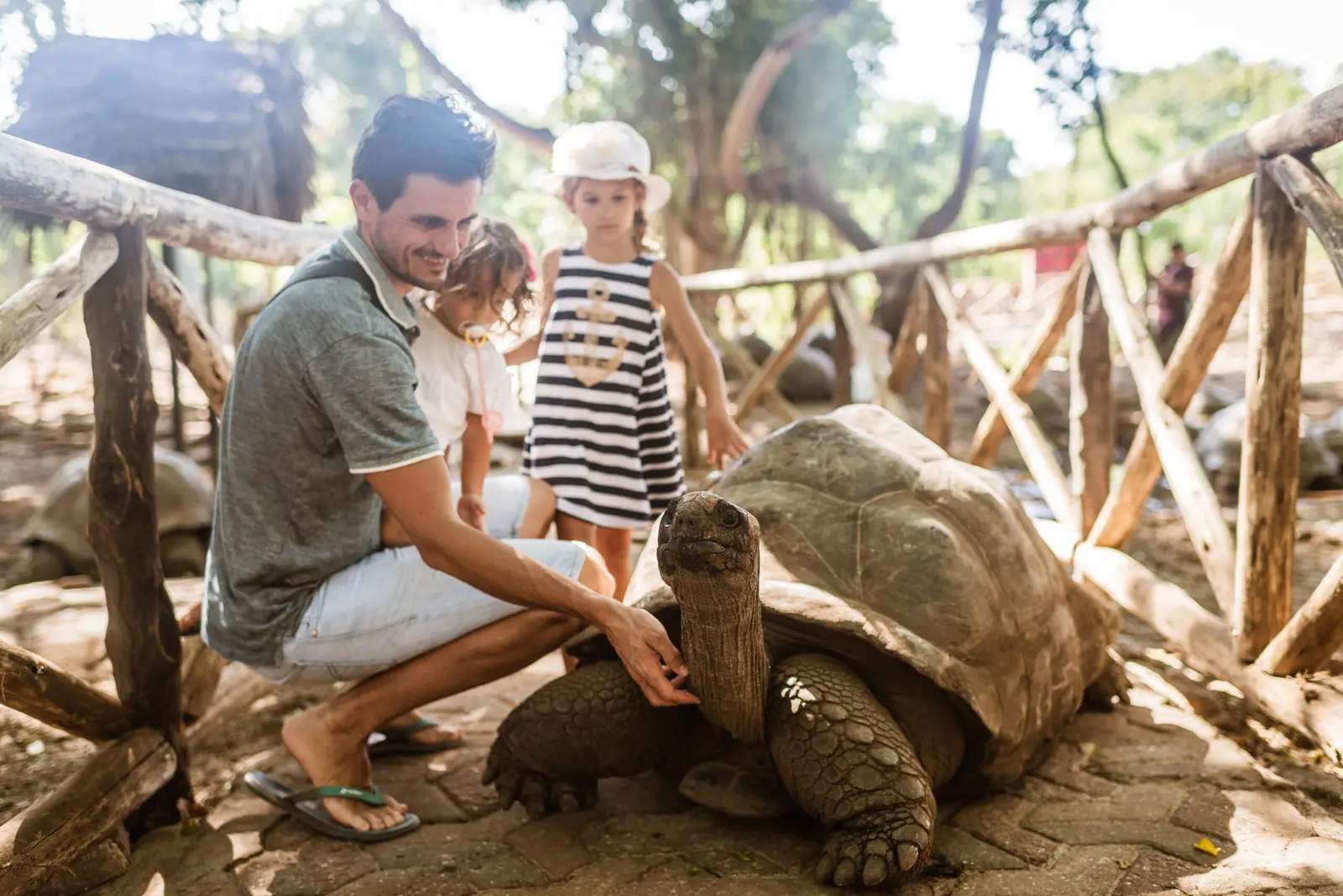 A man and two children petting a giant tortoise inside a rustic wooden enclosure on Prison Island