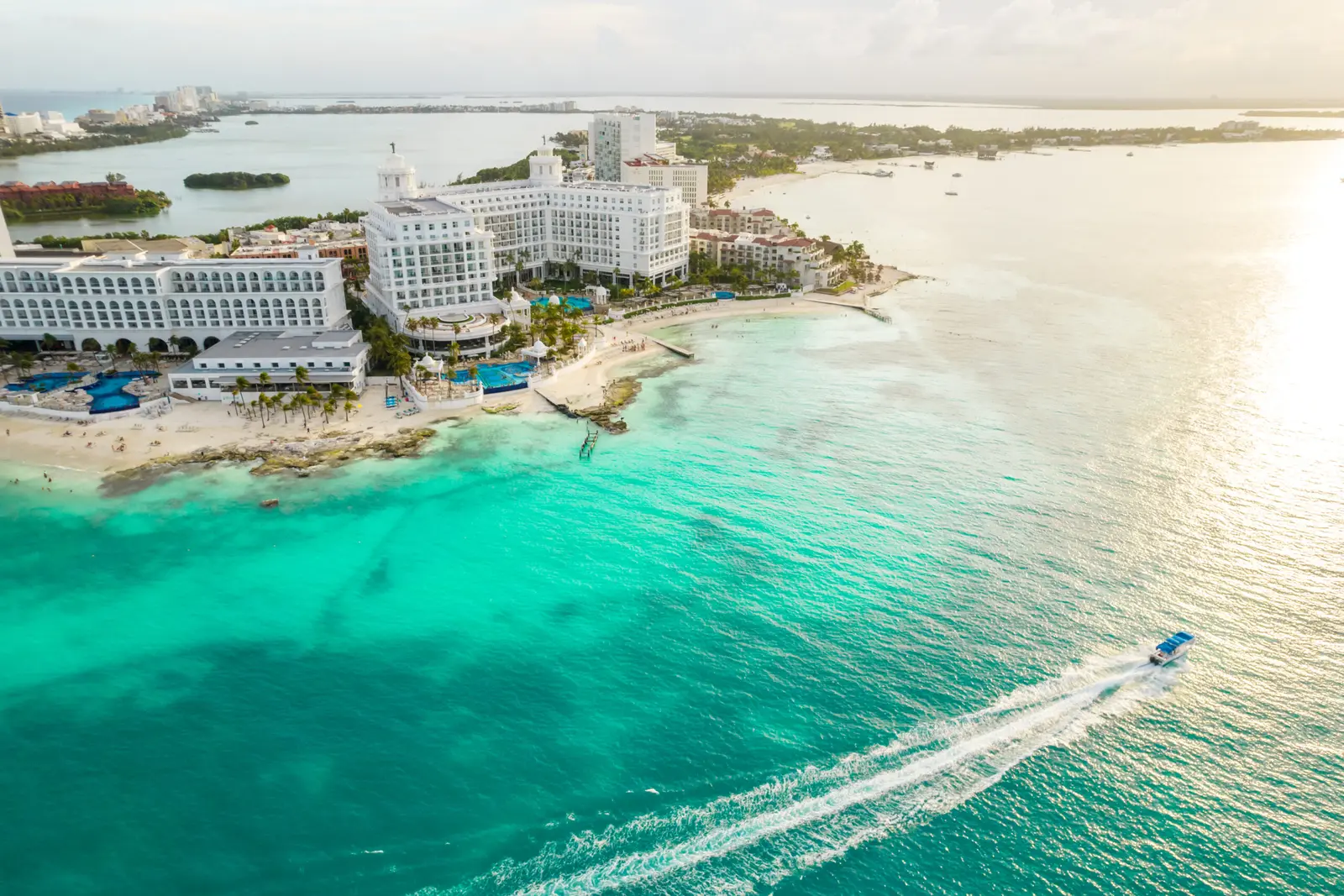 Aerial view of Cancún hotels lining turquoise shallows in late light, a speedboat carving a wake offshore