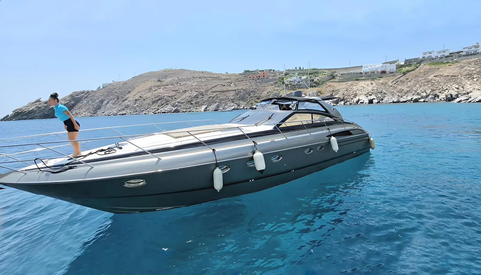 Dark grey motor yacht anchored in calm blue water off the rocky Mykonos coast, a guest on the bow