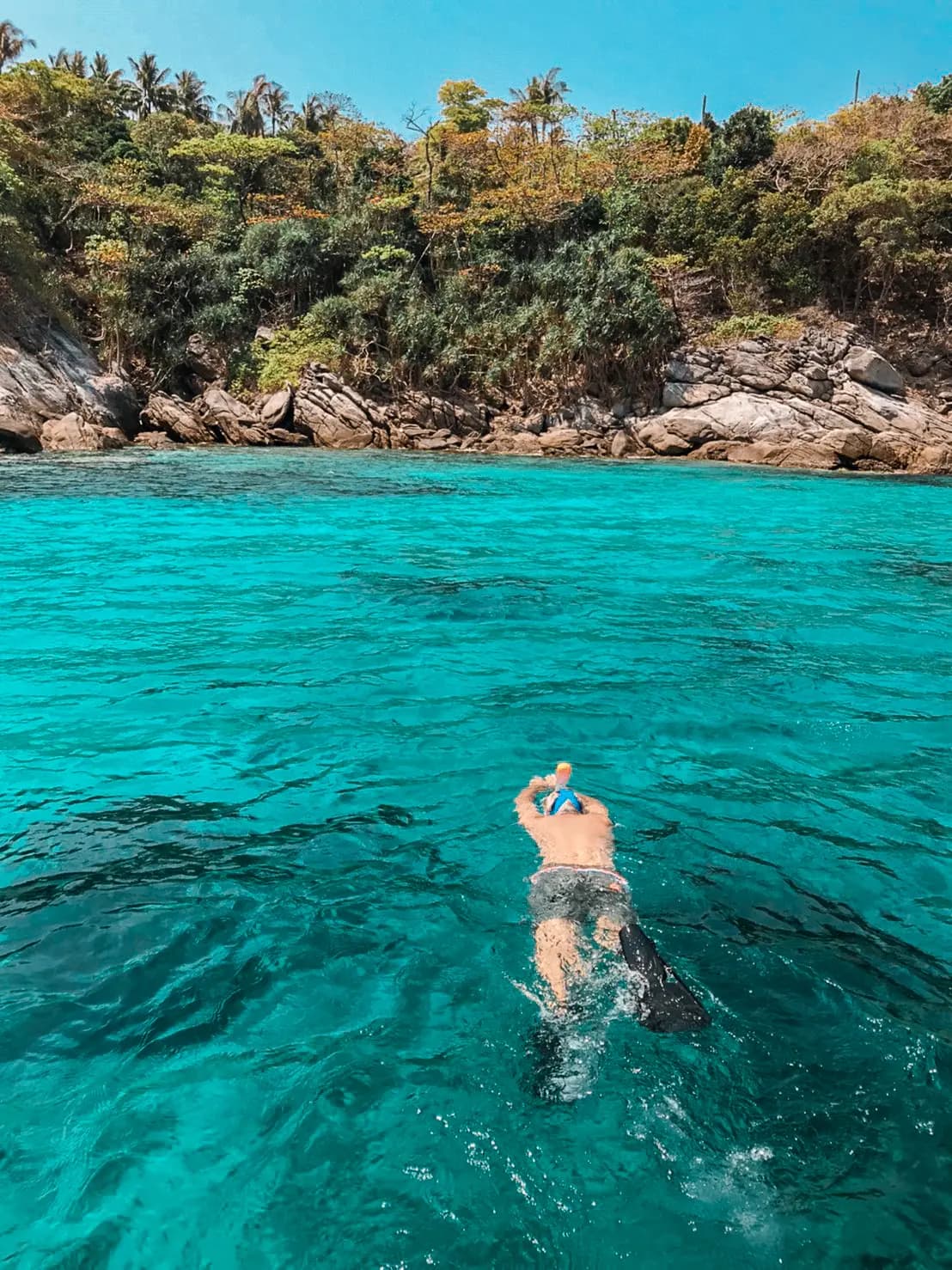 Lone snorkeler swimming toward a rocky, tree-lined shore through clear turquoise water south of Phuket