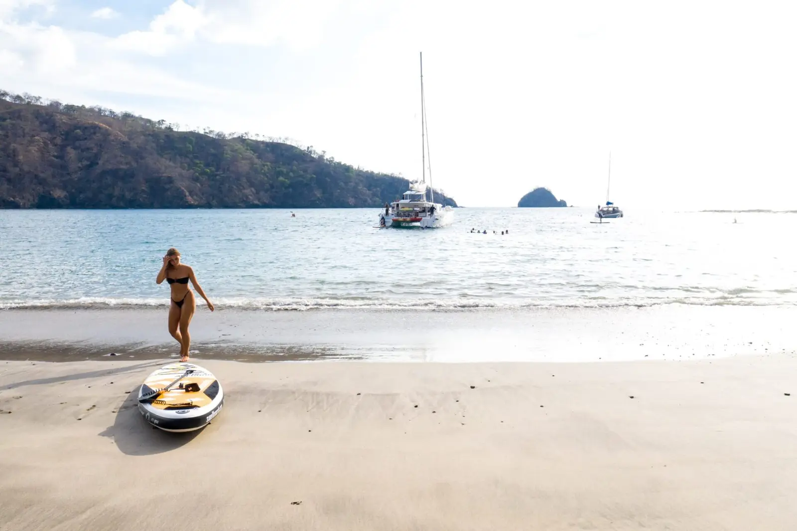 Woman walking a wide Costa Rica beach past a small inflatable, the sailing catamaran anchored offshore
