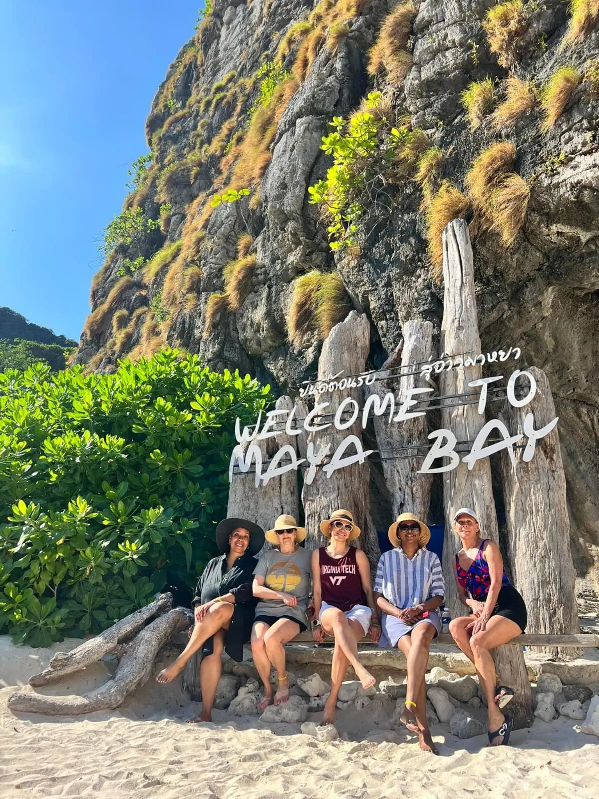 Five guests sitting below the wooden Welcome to Maya Bay sign at the foot of a limestone cliff