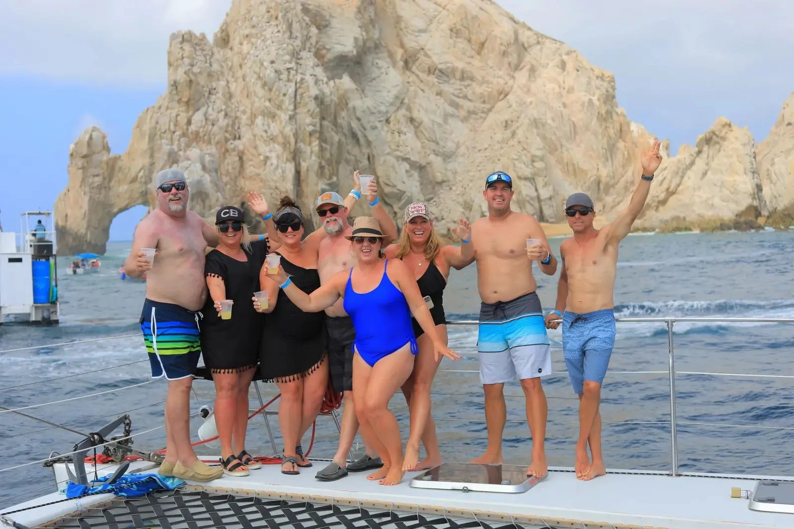 Group of guests in swimwear raising drinks on a catamaran deck with the Cabo San Lucas arch behind